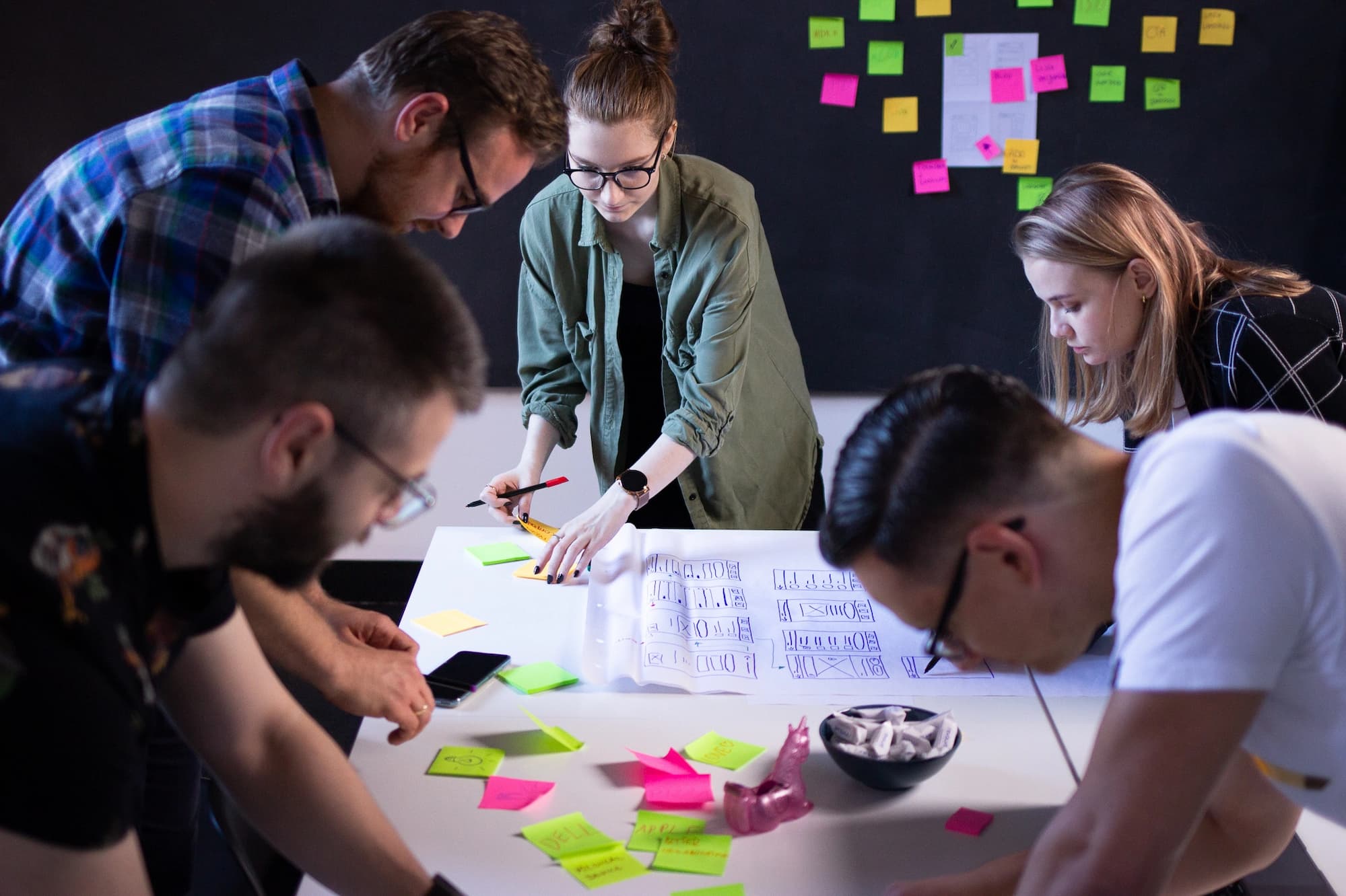 Design workshops at Revolve Healthcare office Design workshops – five people standing over a table, with many sticky notes around and one woman drawing a user interface on a big sheet of paper